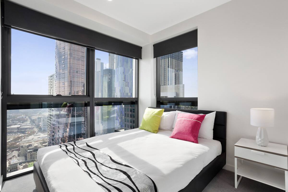 Quarto de hotel moderno com uma cama grande, uma mesinha de cabeceira e vista para a paisagem urbana através de janelas do chão ao teto.