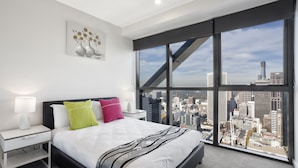 Habitación de hotel moderna con una cama grande, mesitas de noche blancas y vistas a la ciudad a través de ventanas de gran altura.