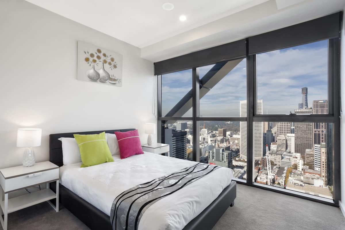Quarto de hotel moderno com uma cama grande, mesinhas de cabeceira brancas e vista para a cidade através de janelas do chão ao teto.
