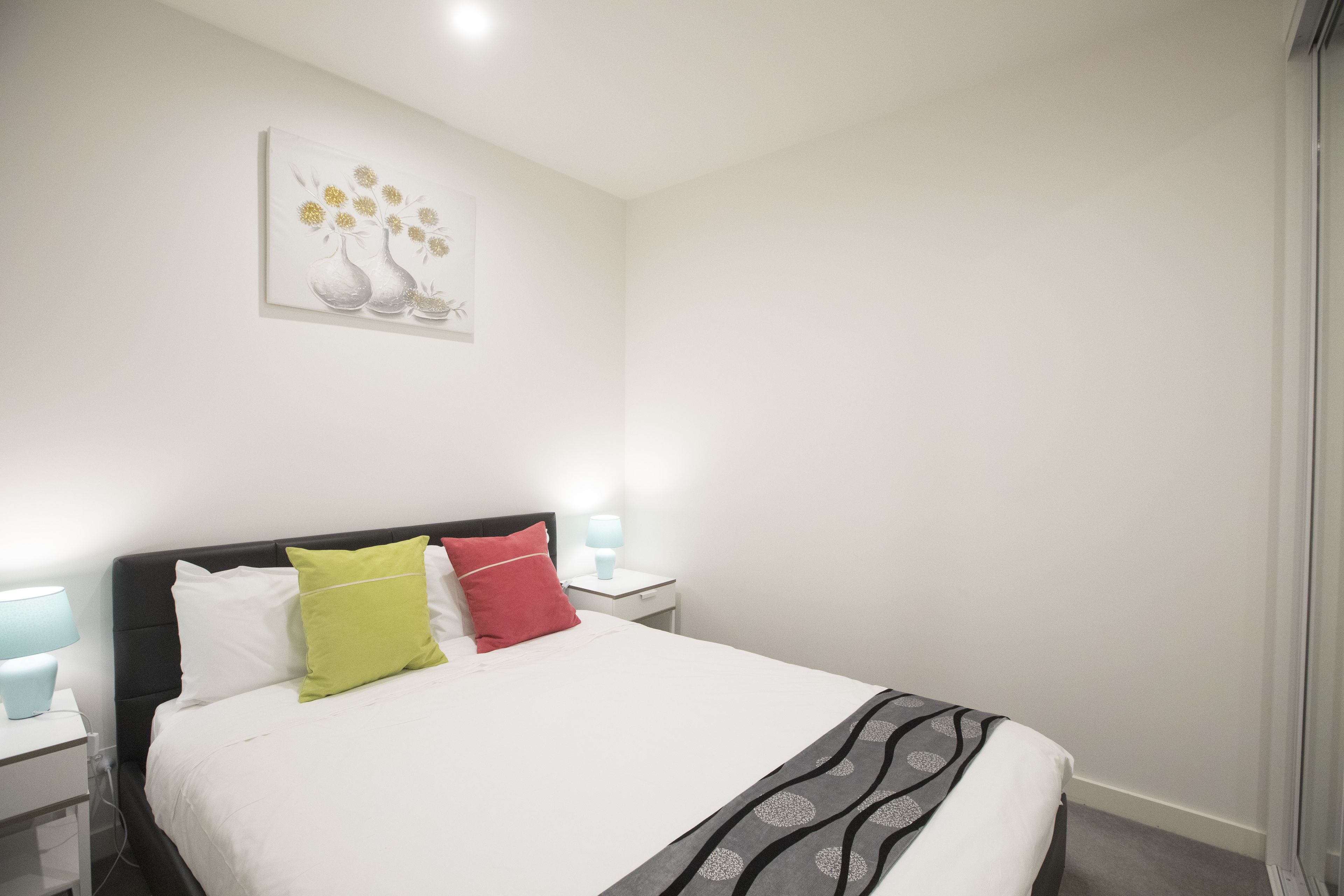 A neatly made bed with white bedding and a gray patterned throw. There is a nightstand with a lamp and a small table beside the bed. A painting of vases with flowers hangs on the wall above the bed.
