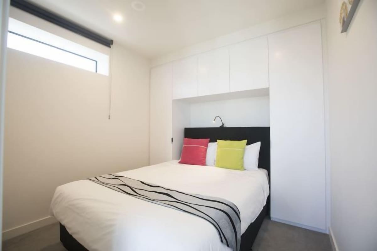 Un dormitorio con cama, paredes blancas, una ventana y cabecera con lámpara.