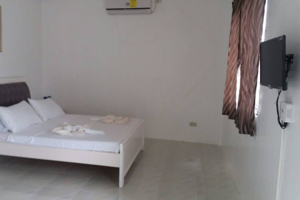 Uma cama branca com travesseiros e um cobertor, um ar condicionado fixado na parede e uma televisão na parede.