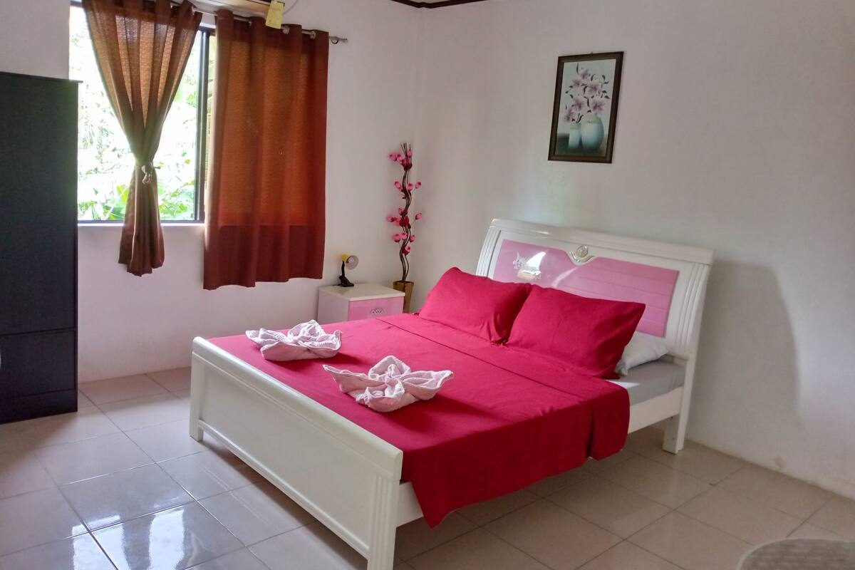 Cama bem arrumada com colcha e almofadas vermelhas, mesa de cabeceira com abajur e janela com cortinas.