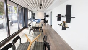 Un espacio de oficina moderno con una mesa larga, sillas y un divisor de vidrio. Hay ventanas amplias y un techo blanco con luminarias lineales.