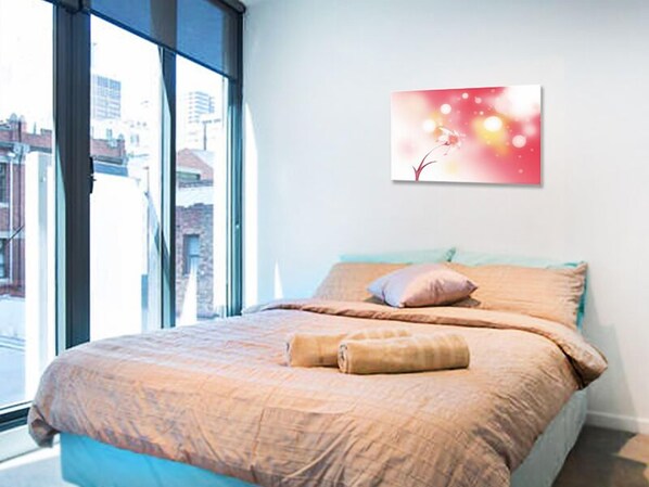 A bedroom with a large window, a bed with beige bedding, and a wall-mounted picture.