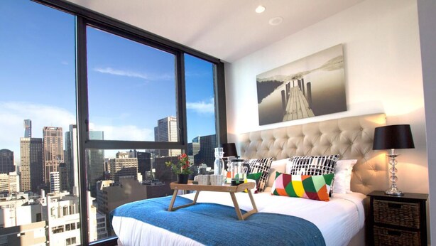 Un dormitorio moderno con un ventanal grande, vista a la ciudad, cabecera capitonada y mesita de noche con lámpara.