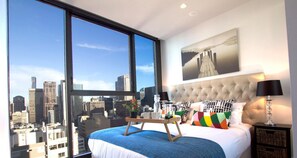 Un dormitorio moderno con un ventanal grande, vista a la ciudad, cabecera capitonada y mesita de noche con lámpara.