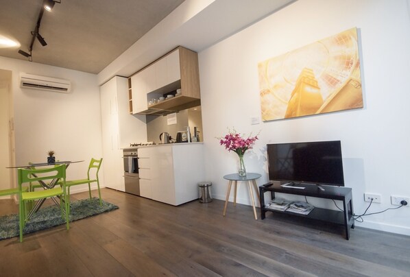 Una cocina moderna con armarios blancos, una zona de comedor con sillas verdes, un televisor sobre un mueble bajo y un cuadro en la pared.