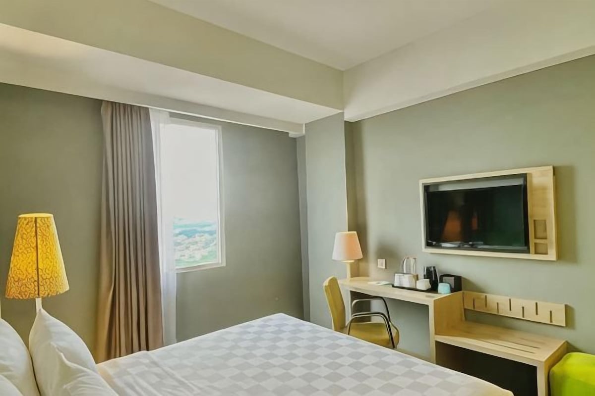 A hotel room with a bed, a desk with a chair, a television, and a window with curtains.