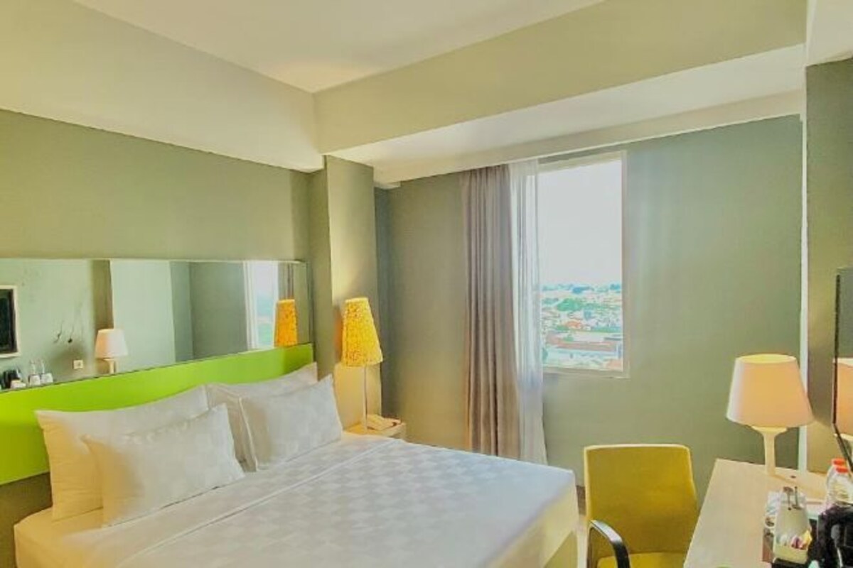 Habitación de hotel con cama, escritorio, silla, lámpara, ventana con cortinas y vista al exterior.