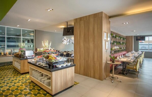 A modern hotel dining area with a buffet setup, a long communal table, and a seating area with a wooden accent wall.