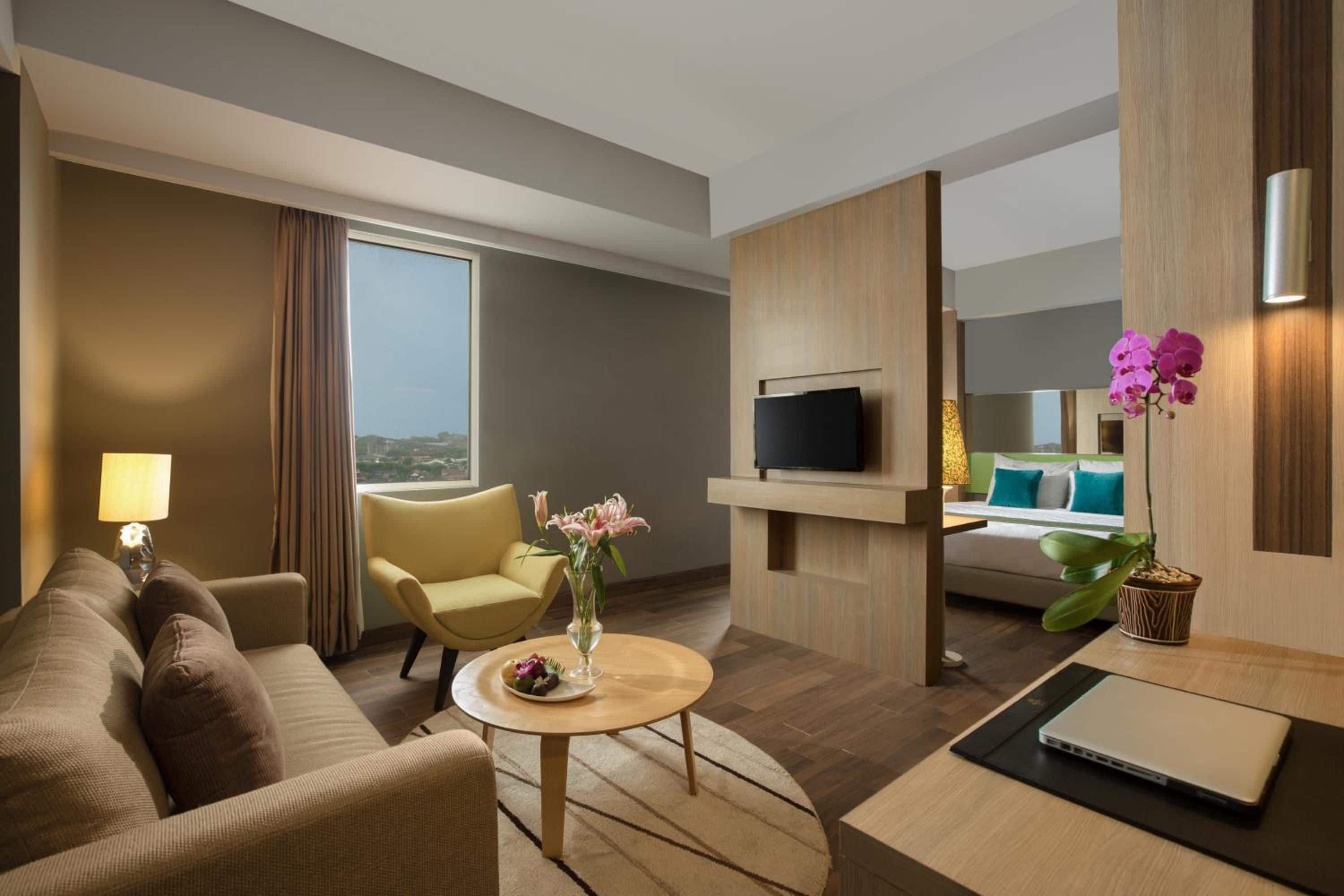 A hotel room with a sofa, armchair, coffee table, TV, and a bed with a view.