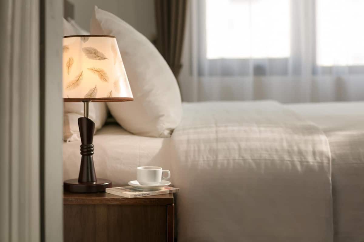 Una cama bien tendida con edredón blanco, una mesita de noche con lámpara, una taza y un libro.