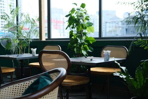 Un espace repas avec des tables et des chaises, de grandes fenêtres et des plantes d’intérieur.