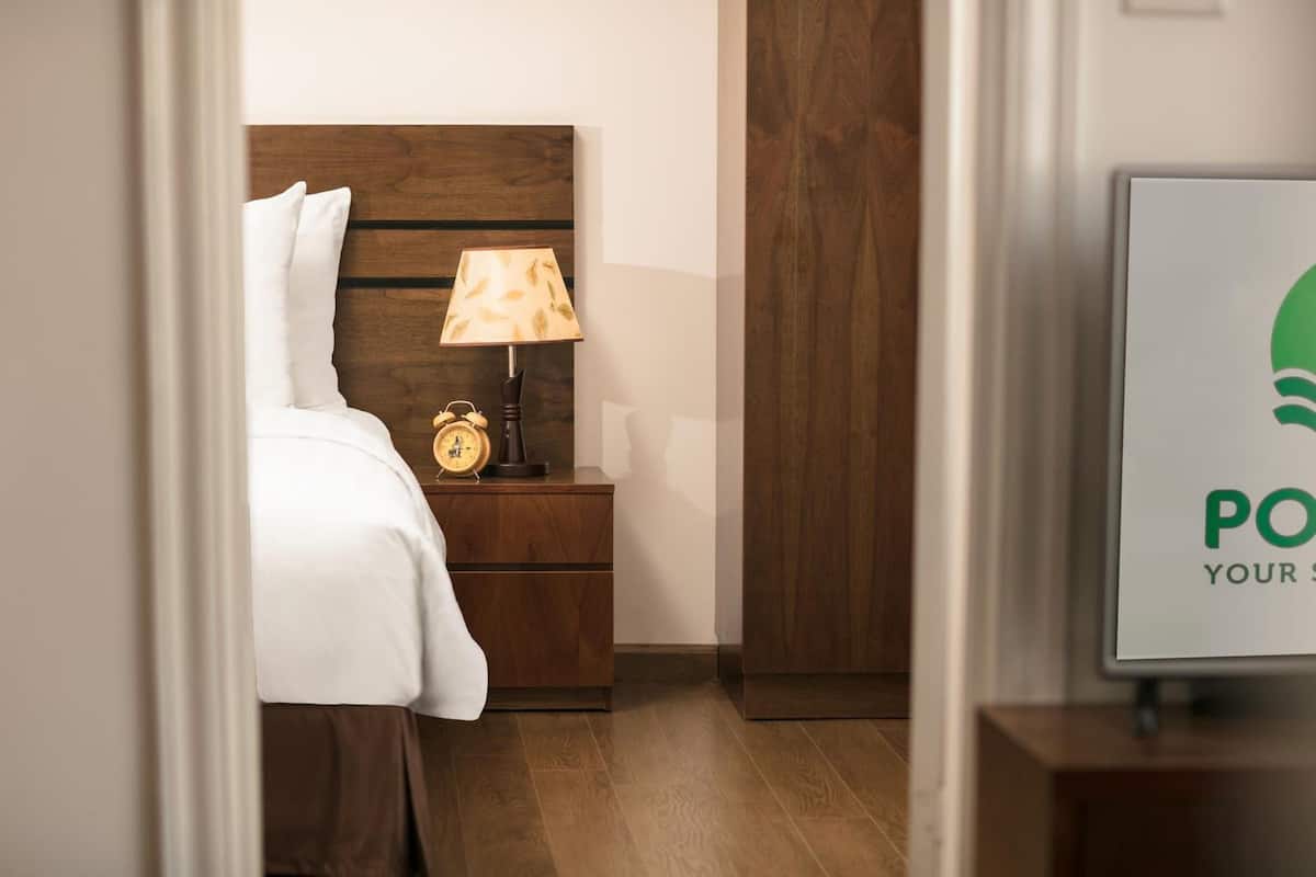 Habitación de hotel con cabecera de madera, mesita de noche con lámpara y despertador, y un cartel enmarcado en la pared.
