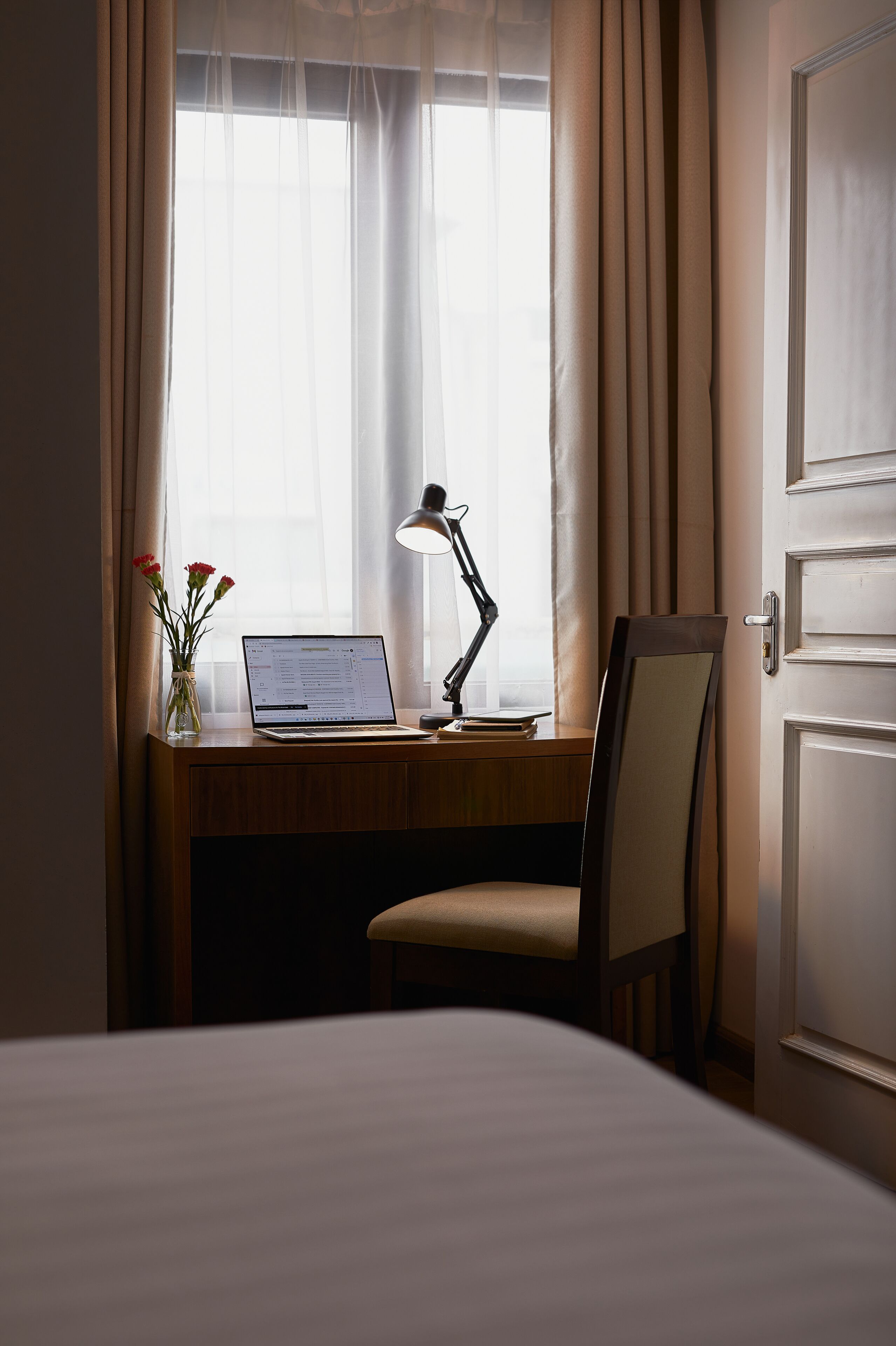 A hotel room with a wooden desk, a laptop, a desk lamp, and a vase with flowers.