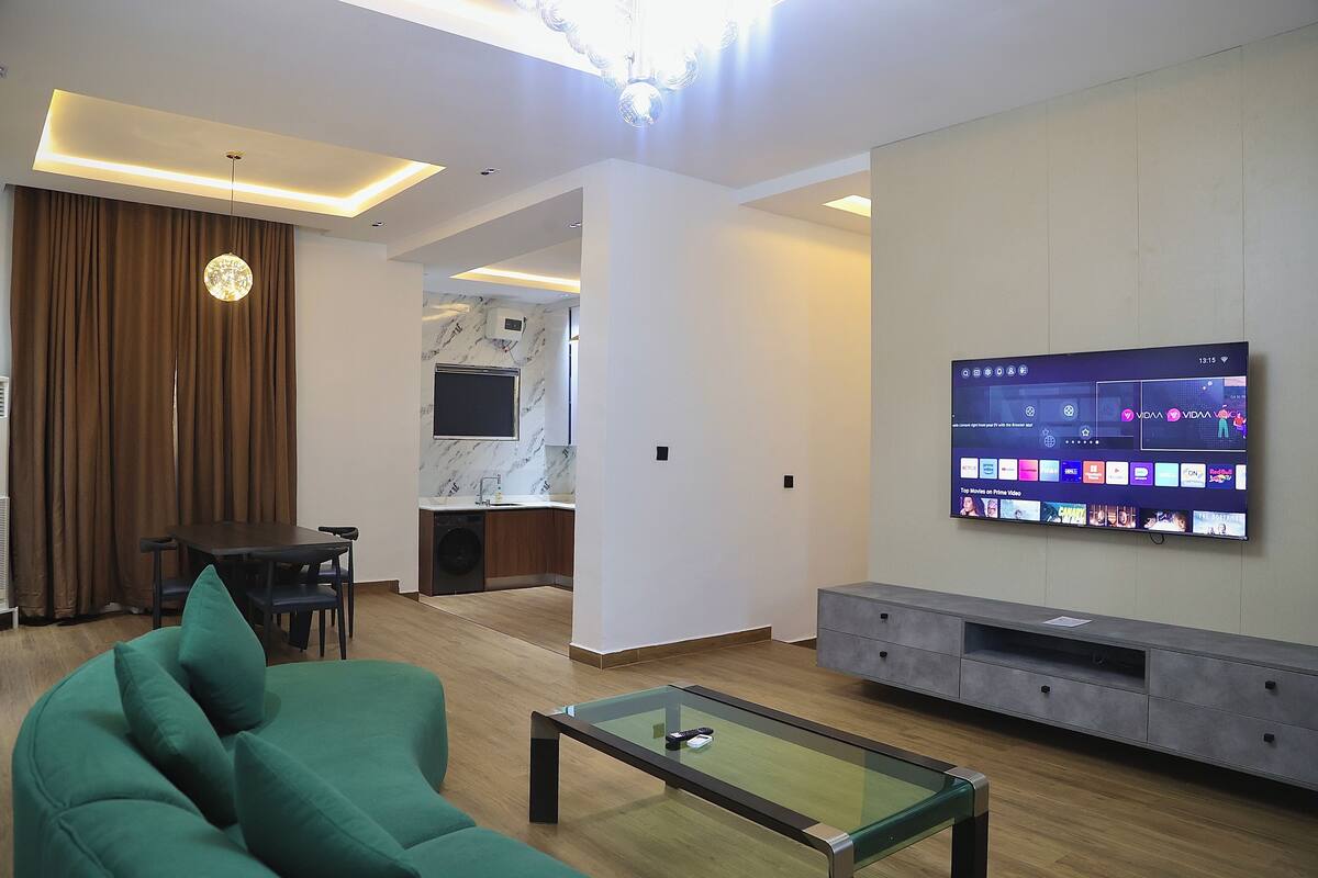 A modern living room with a green sofa, a glass coffee table, a flat-screen TV mounted on the wall, and a kitchen area in the background.