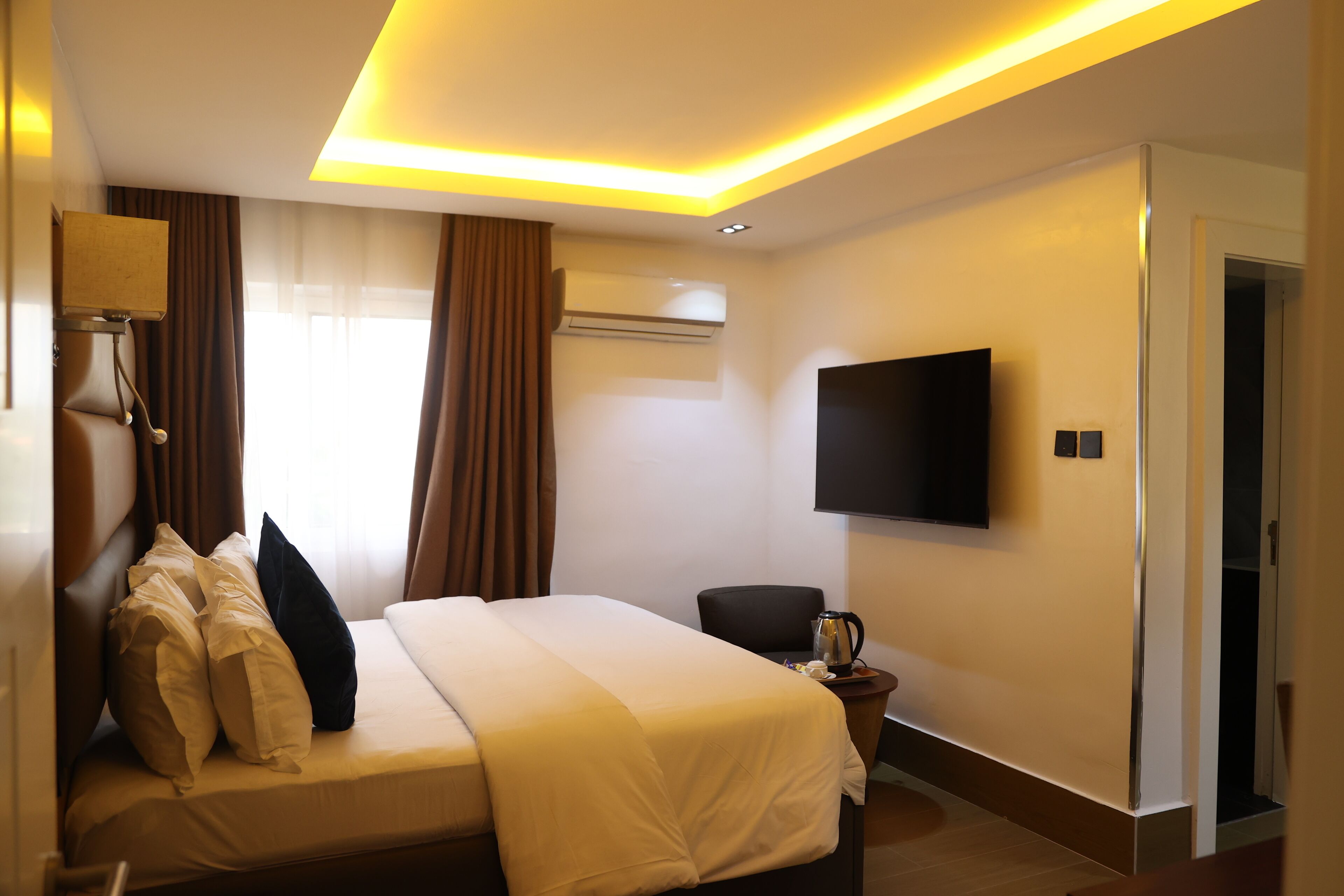 A hotel room with a bed, a flat-screen TV, an air conditioning unit, and a coffee maker on a side table.