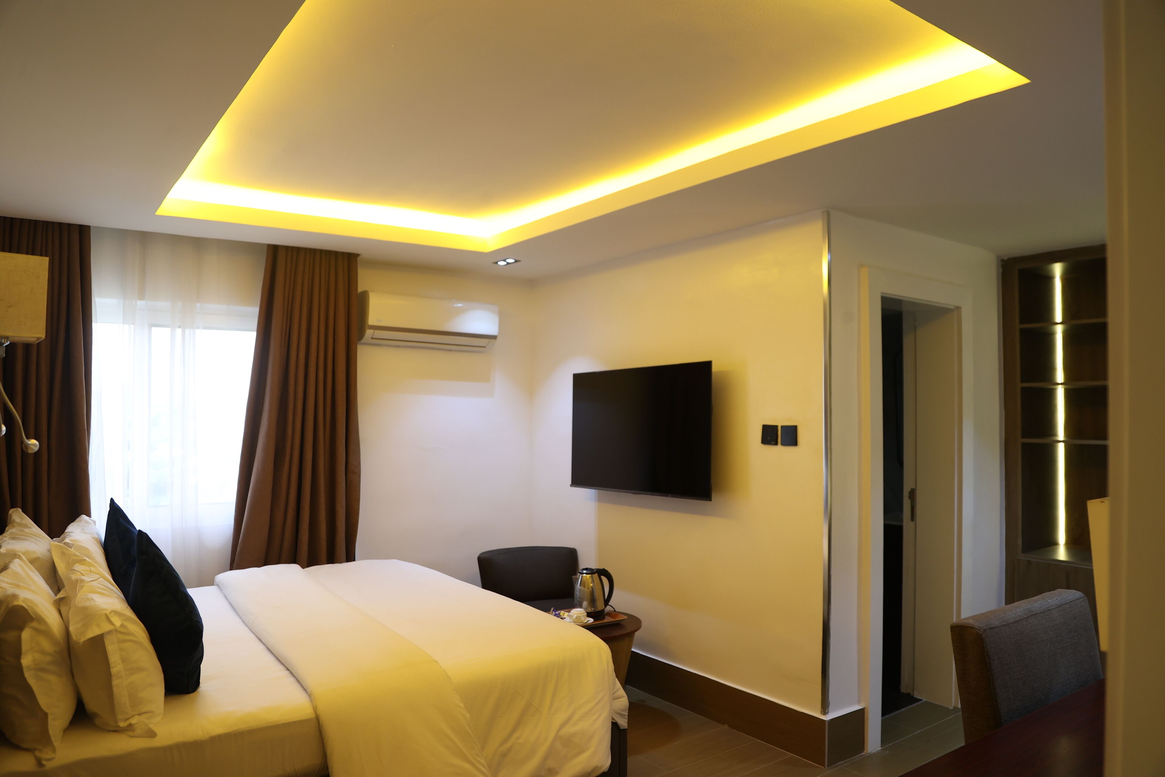 A hotel room with a bed, a TV mounted on the wall, a coffee maker on a side table, and a door leading to another room.