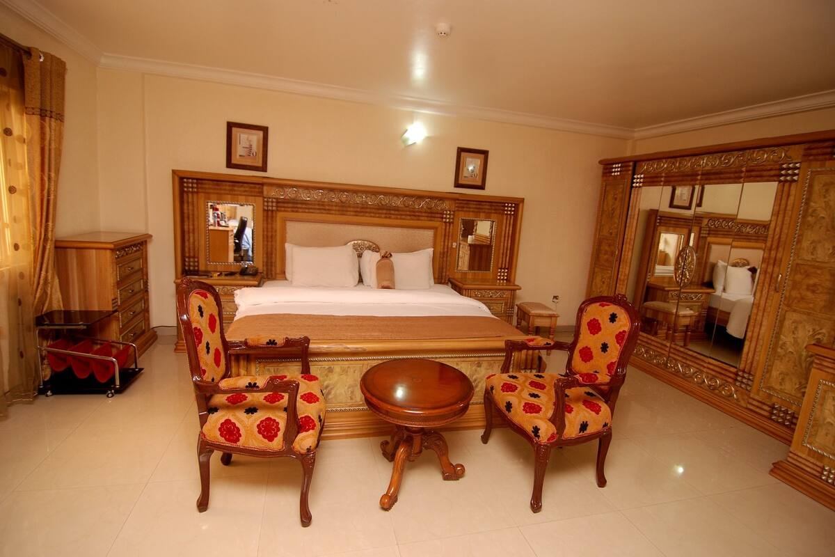 Un dormitorio amplio con una cama grande, dos sillas ornamentales, una mesa redonda y un espejo de gran tamaño.