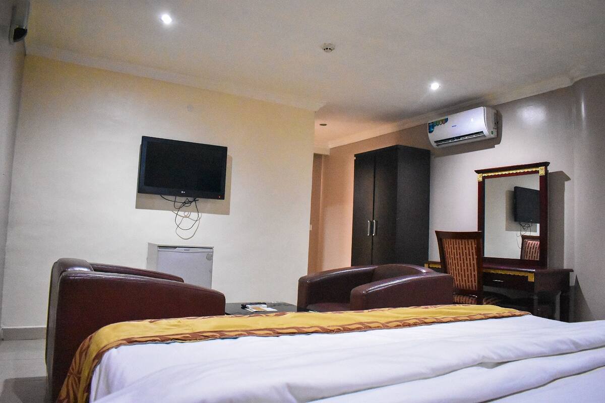 A hotel room with a bed, two leather chairs, a desk, a flat-screen TV, an air conditioning unit, a wardrobe, and a mirror.