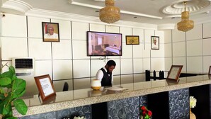 A hotel reception area with a reception desk, a television mounted on the wall, framed certificates, and a potted plant. - Laps New World Hotel (Abuja)