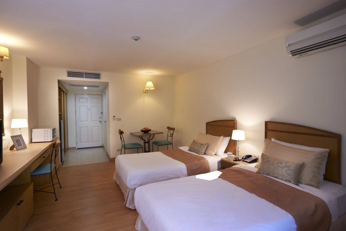 Habitación de hotel con dos camas, un escritorio con silla, una mesita con sillas, una lámpara y un equipo de aire acondicionado.