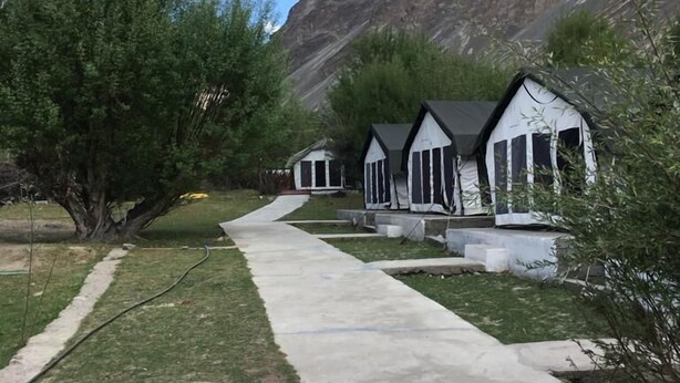 A pathway leading to a row of white cabins with black trim, set against a mountainous backdrop. - Nubra Summer Camp (Leh)