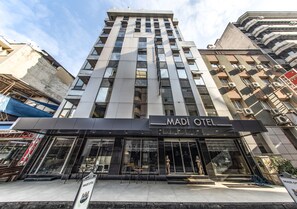 Een modern hotel met een opvallend bord waarop 'MADI OTEL' staat. Het heeft een ruime entree met glazen deuren en een zitgedeelte buiten.