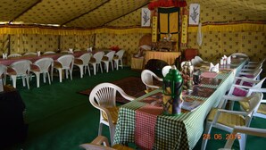 Un área de comedor con toldo, mesas dispuestas para varios comensales, sillas de plástico blanco y una mesa central con un centro de mesa decorativo.