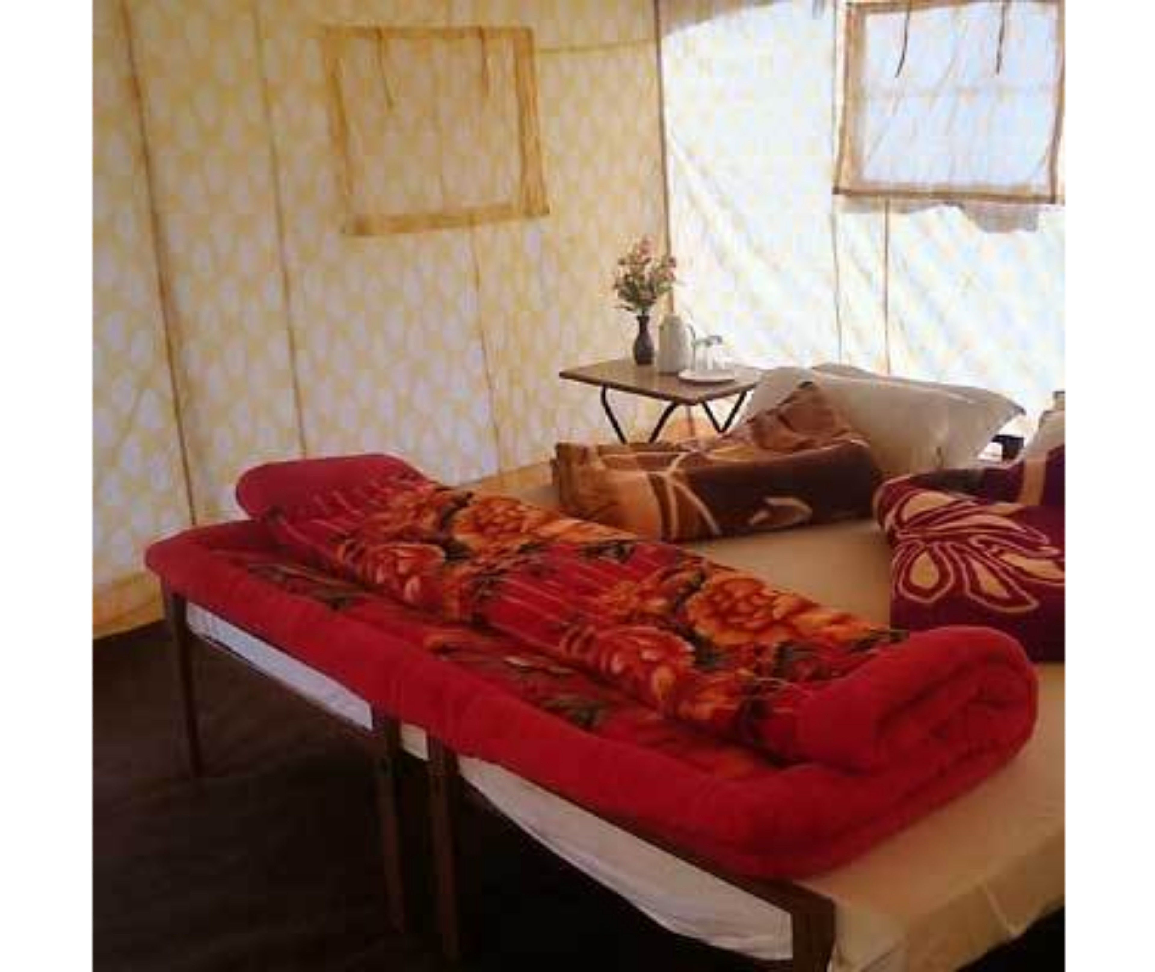 Una cama individual con una colcha roja, una mesita con un florero y una ventana con cortinas.