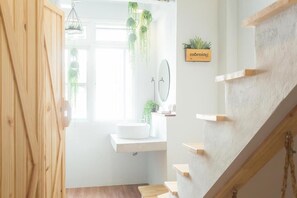A modern bathroom with a wooden door, a white sink, a mirror, and a hanging plant. - Barn and Bed Hostel (Bangkok)