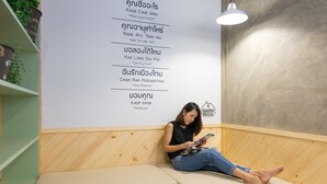 A person sitting on a sofa in a room with a wall displaying Thai phrases and a logo for Barn Bed &. - Barn and Bed Hostel (Bangkok)
