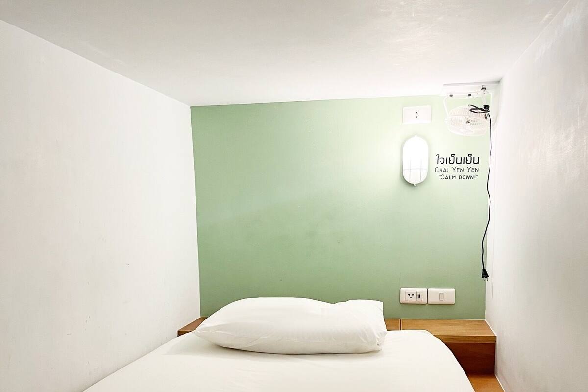 A simple, clean room with a single bed, a nightstand, and a wall-mounted light.