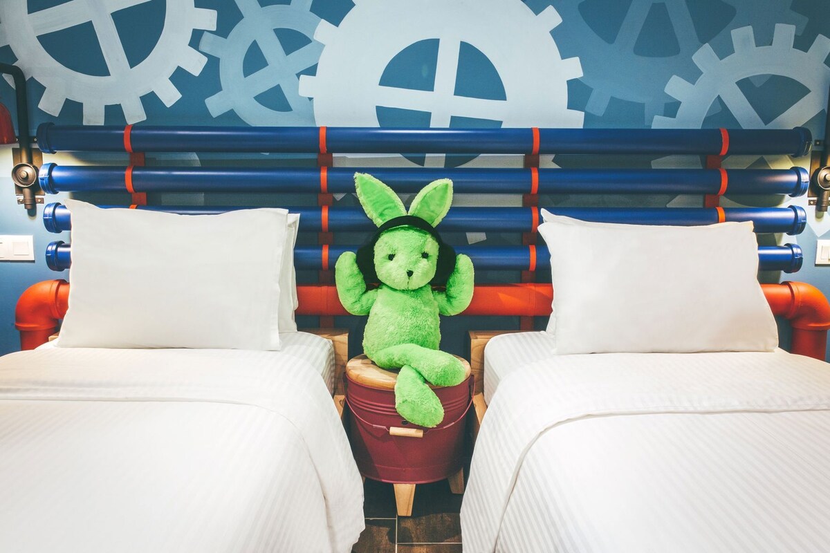 A room with two beds, a green plush toy on a stool, and a wall with large gears.