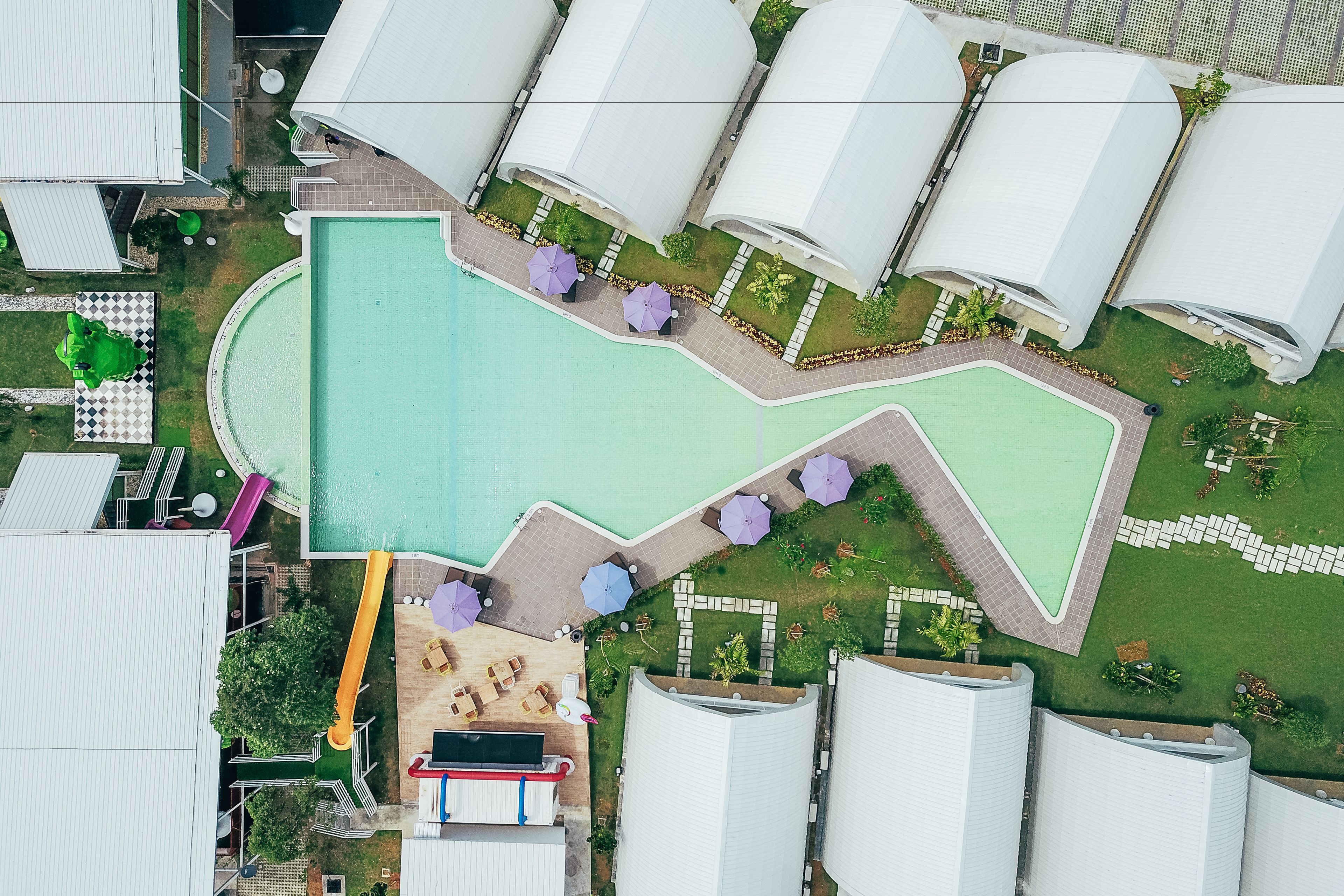 An aerial view of a resort with a large swimming pool, multiple umbrella seating areas, and a children's play area.