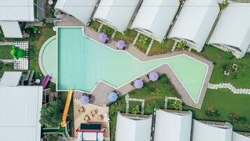 An aerial view of a resort with a large swimming pool, multiple umbrella seating areas, and a children's play area.