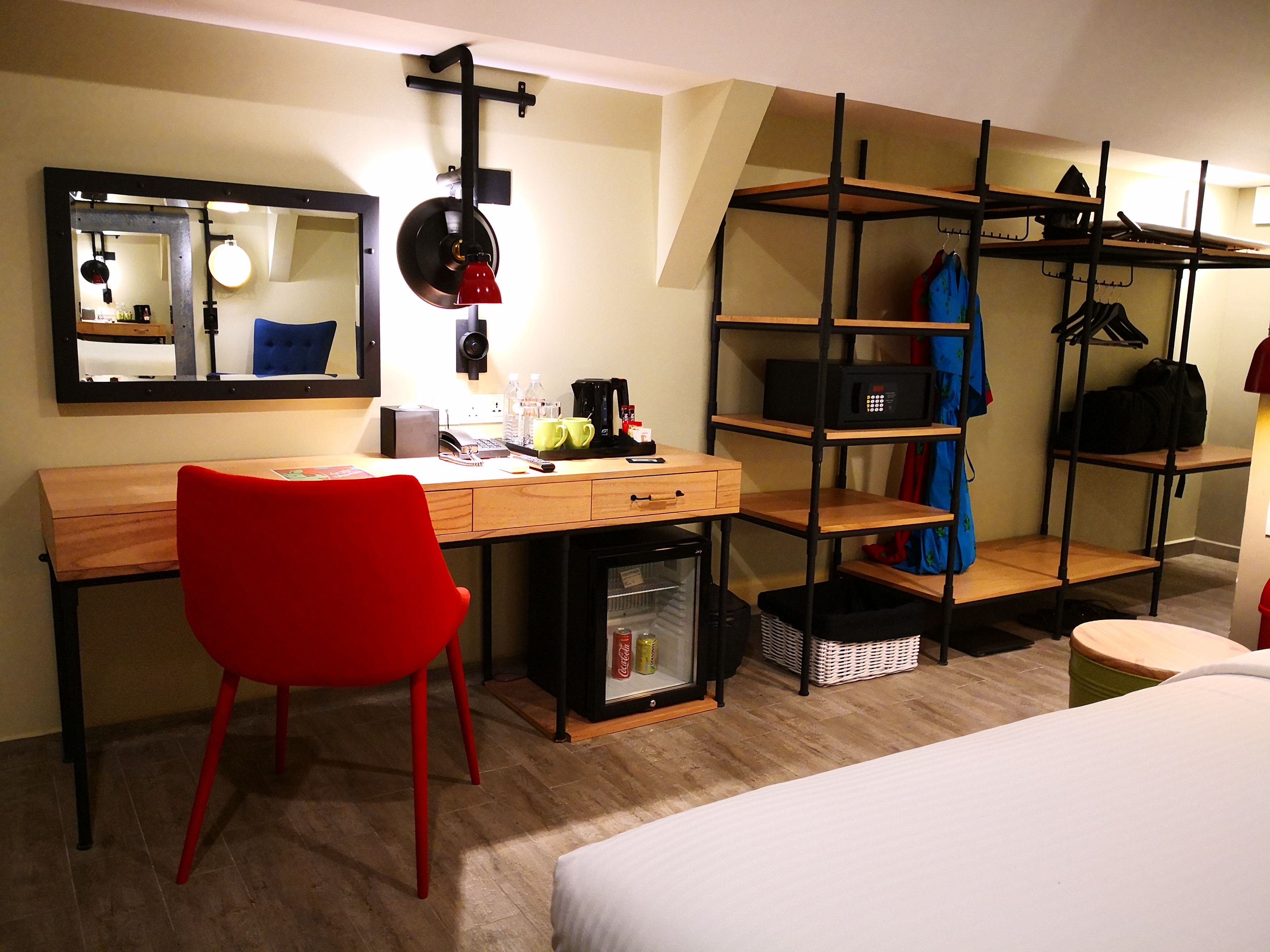 A hotel room with a wooden desk, a red chair, a bed, a wardrobe with shelves, a mirror, and a small refrigerator.