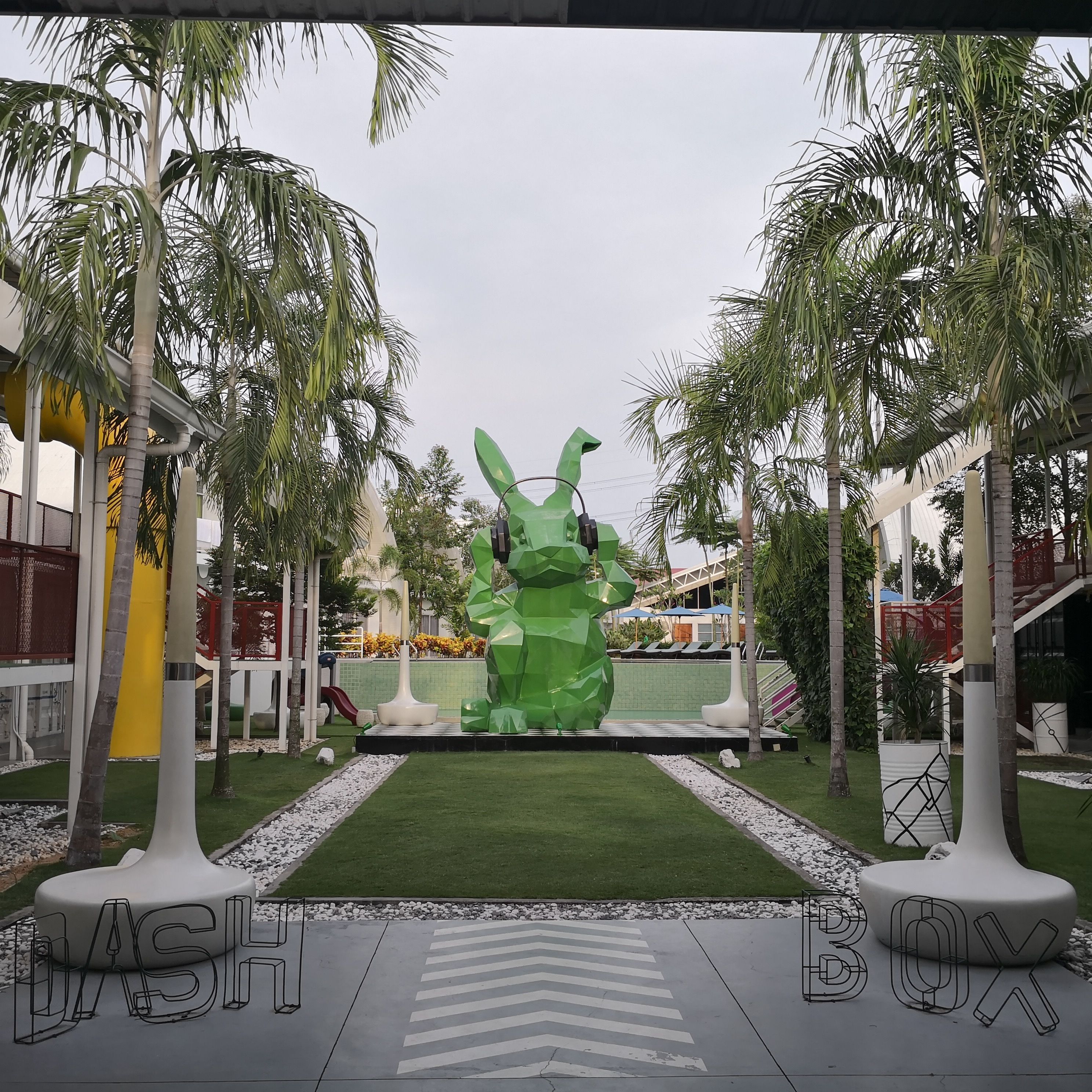 A modern outdoor area with a large green sculpture, palm trees, and a pathway leading to a building with a yellow facade.
