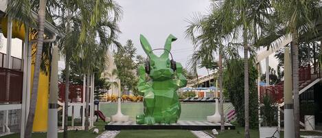 A modern outdoor area with a large green sculpture, palm trees, and a pathway leading to a building with a yellow facade.