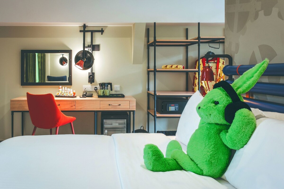 A hotel room with a bed, a desk with a chair, a mirror, a shelf with items, and a green plush toy.