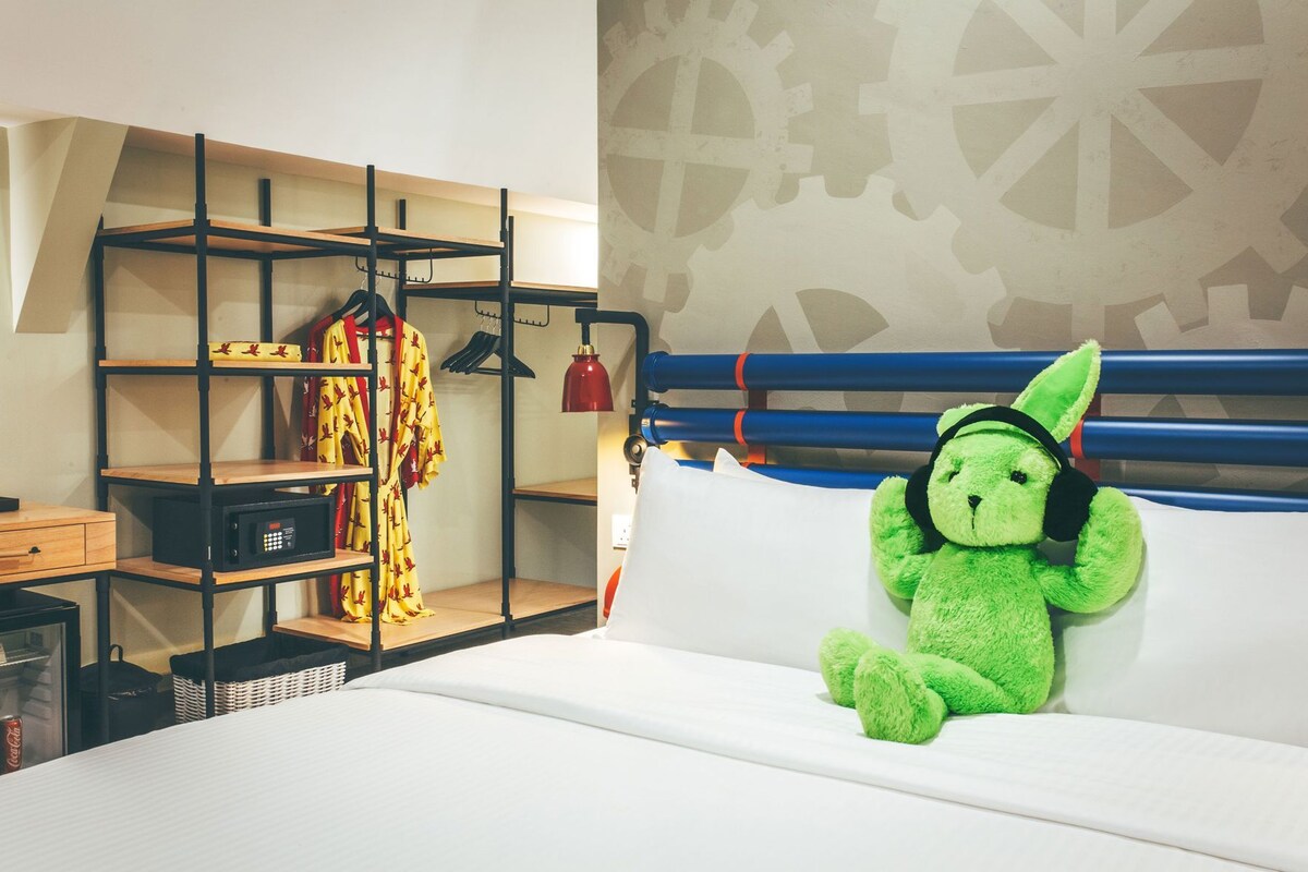 A bunk bed with a green plush toy on the lower bunk, a red lamp, and a clothing rack with a yellow garment.