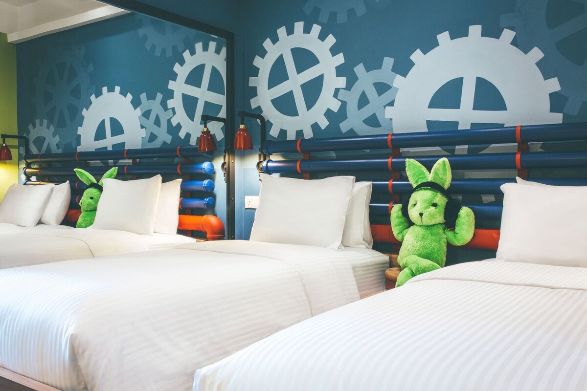 A hotel room with two beds, a wall with large white gears, and a green plush toy on one bed.