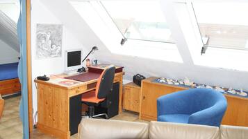 A room with a sloped ceiling, a desk with a computer, a blue chair, a couch, and a window with a view of buildings.