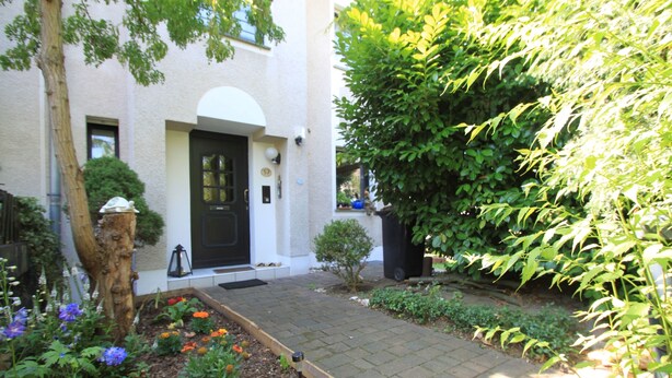 A garden with a paved walkway, a black door, and a small tree. - B&B am See - Privatzimmer (Cologne)