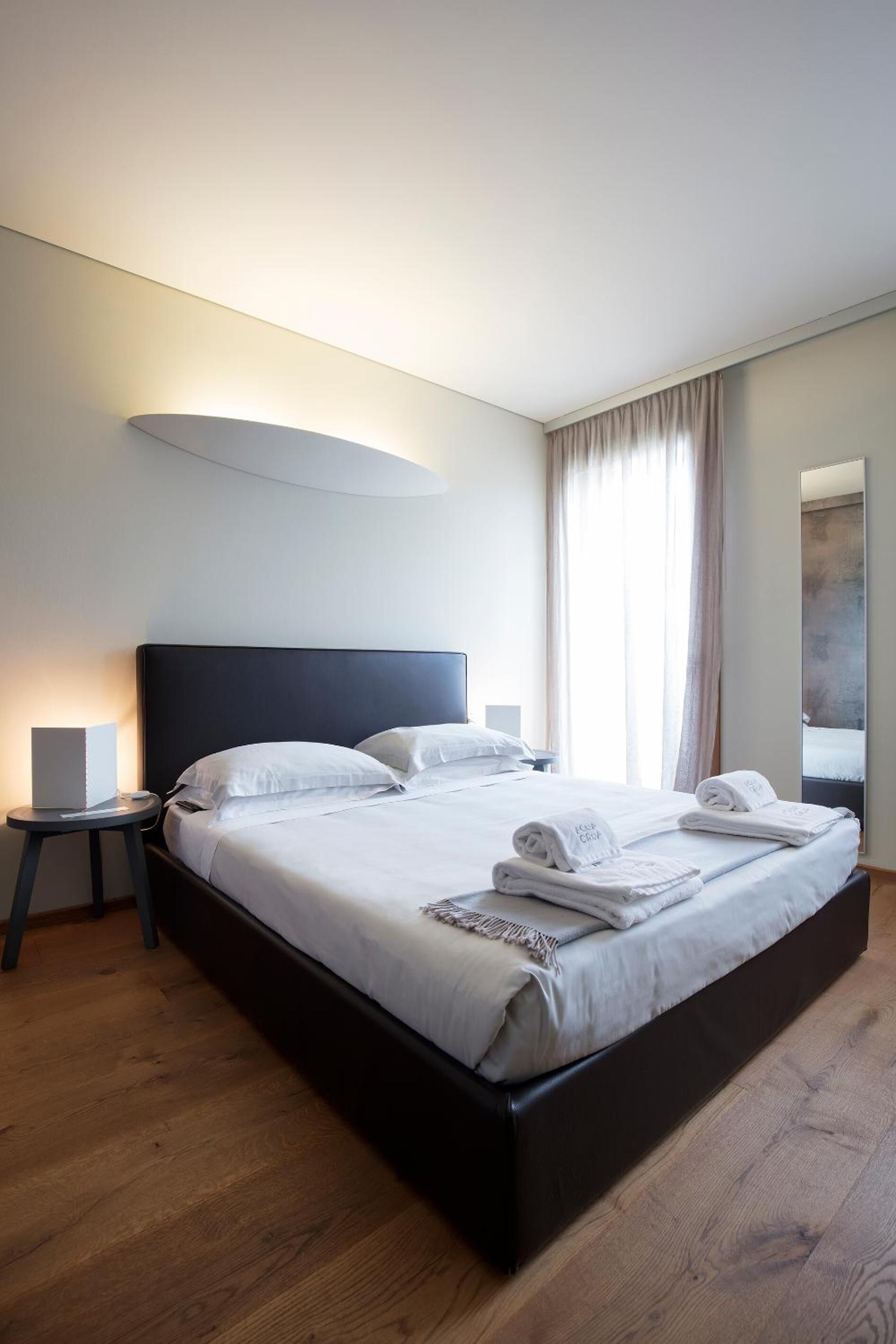 A modern hotel room with a large bed, a bedside table with a lamp, and a view of the city through the window.