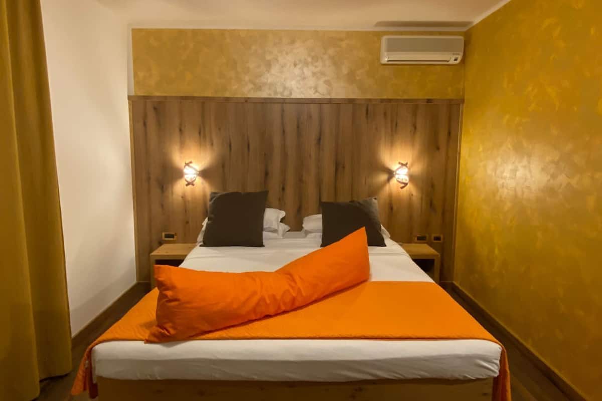 A bedroom with a wooden headboard, two bedside lamps, a bed with white and orange bedding, and an air conditioning unit on the wall.