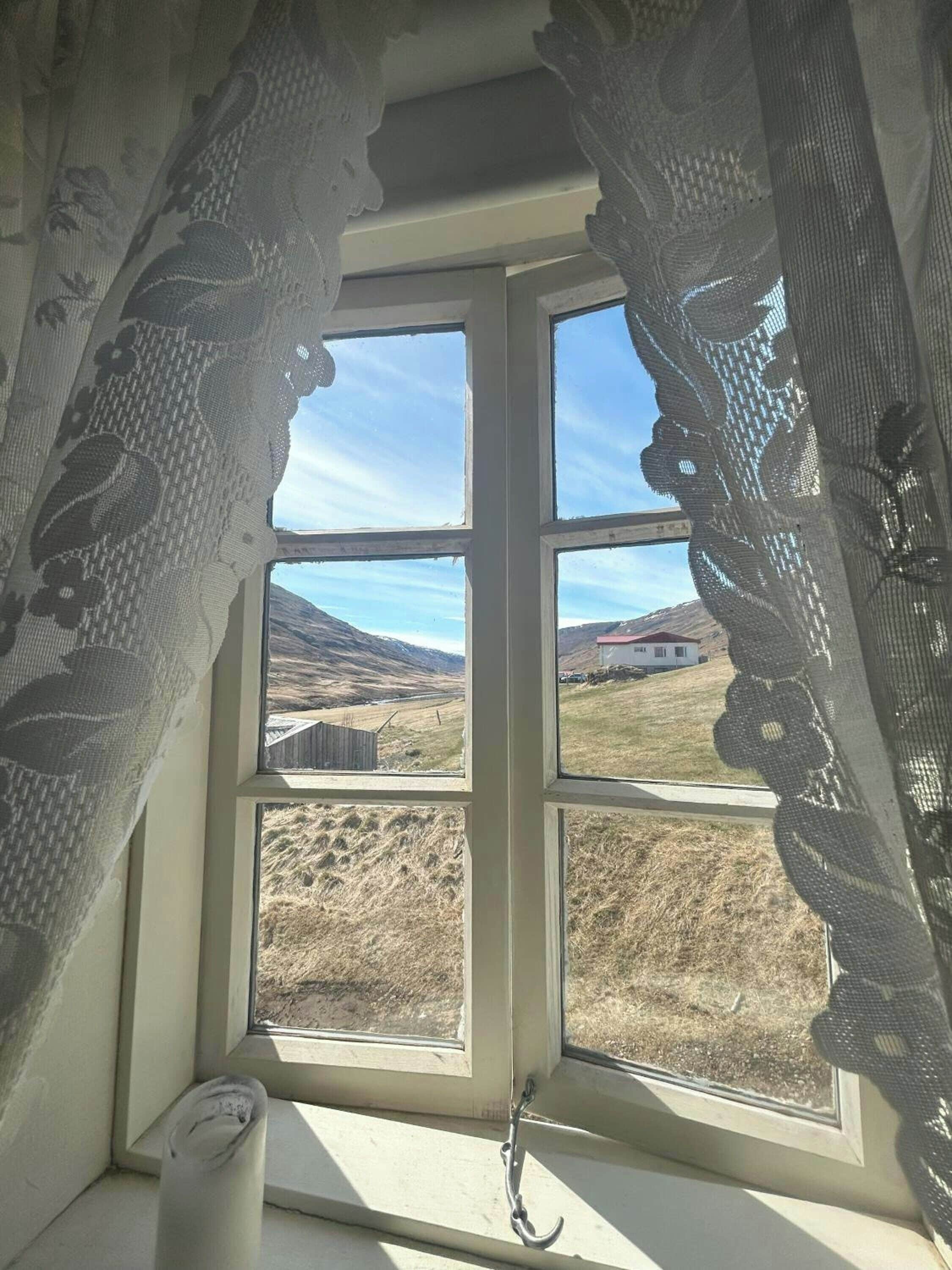 Ein Fenster mit Spitzenvorhängen, Blick auf eine ländliche Landschaft und eine Holzstruktur in der Ferne.