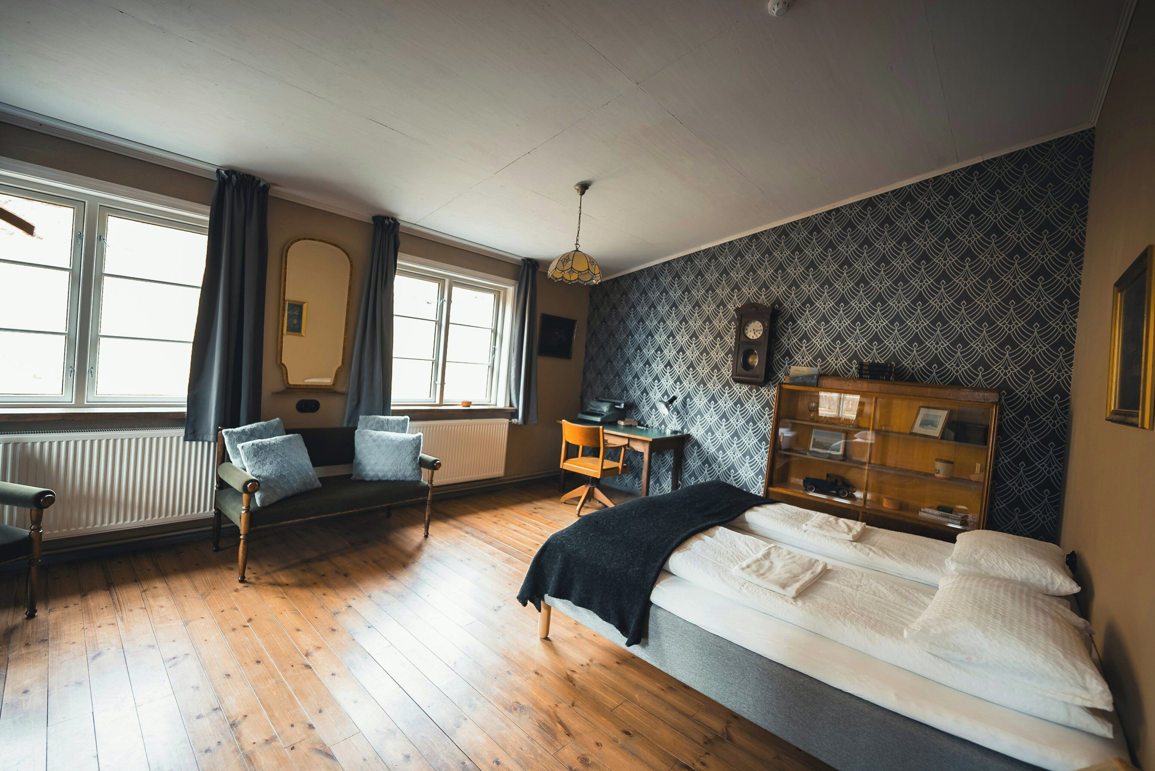 A room with a bed, a desk, a chair, a radiator, and a window with curtains.