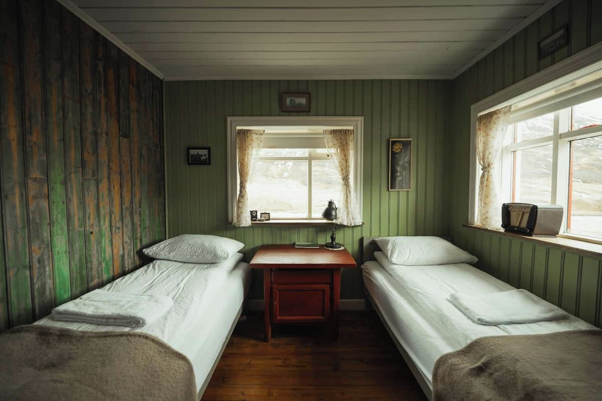 A room with two single beds, a small table, a window with curtains, and a radiator.